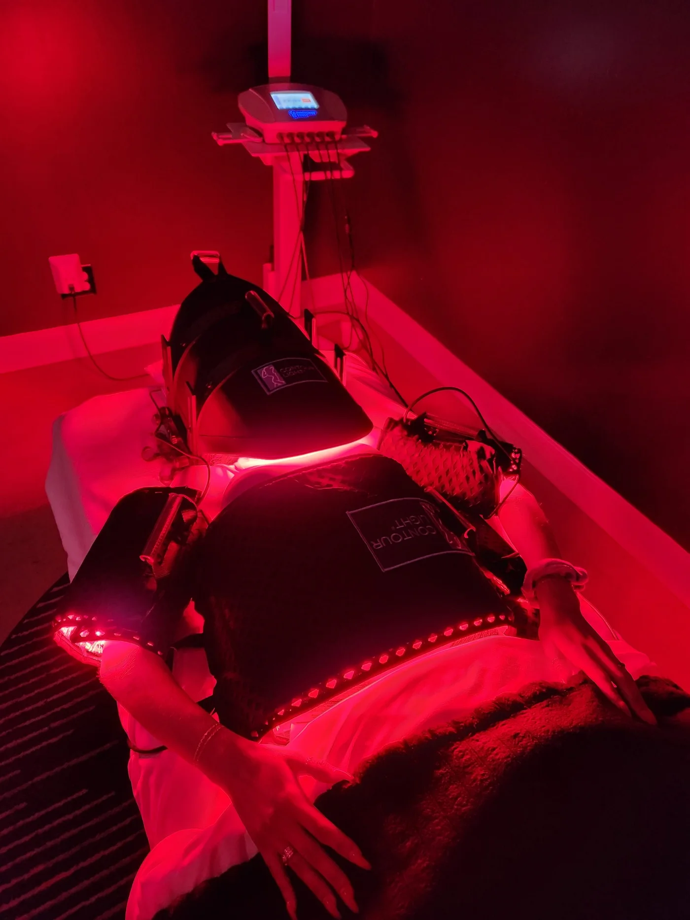 A client resting on a treatment table under Contour Light pads glowing red, the system’s control unit behind her